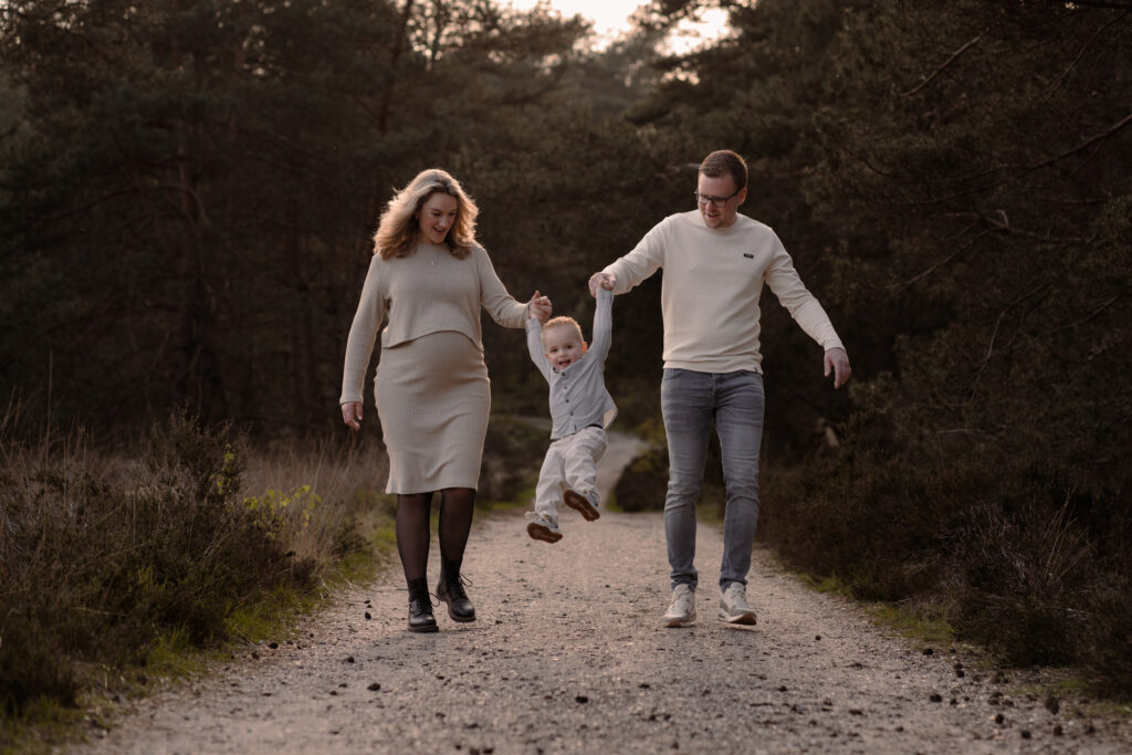 Zwangere vrouw en partner en eerste zoontje tijdens een zwangerschapshoot in de natuur wandelend over een pad door het bos