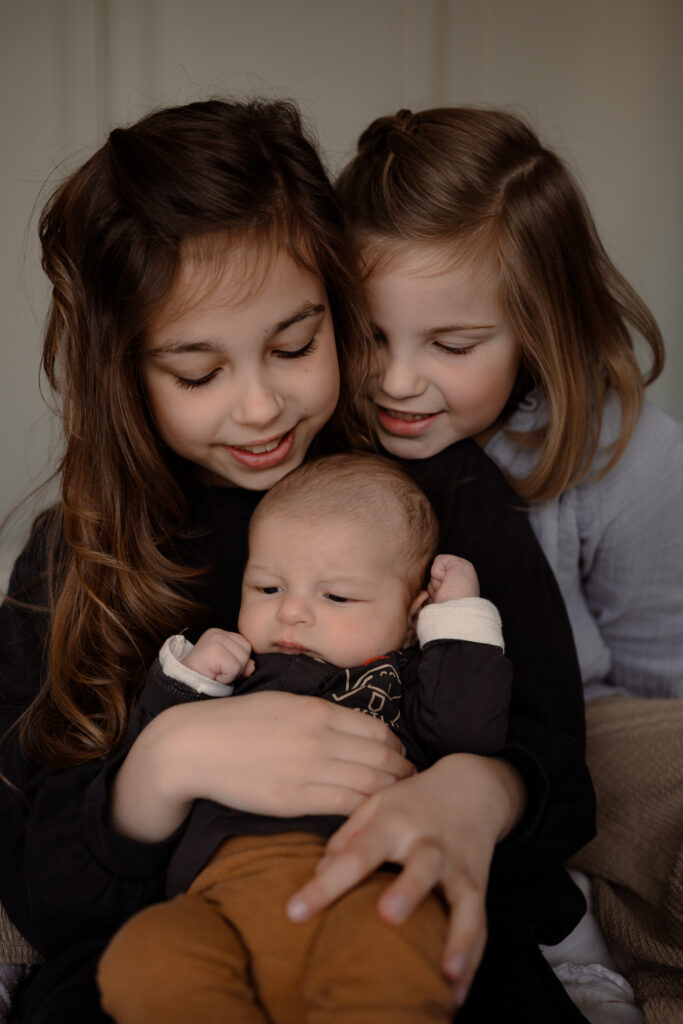 Baby broertje zit bij grote zus op schoot en de andere zus kijkt over haar schouder mee tijdens een newbornshoot aan huis